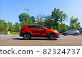 A red new car is driving along the road, standing out in the general traffic. A park with green trees, road signs, and a dividing strip with posts In the background. Driving school concept 82324737