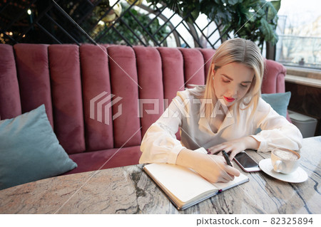 Dreamy young woman writing down future plans and goals in own diary resting in cozy cafeteria Dreamy young woman writing down future plans and goals in own diary resting in cozy cafeteria 82325894