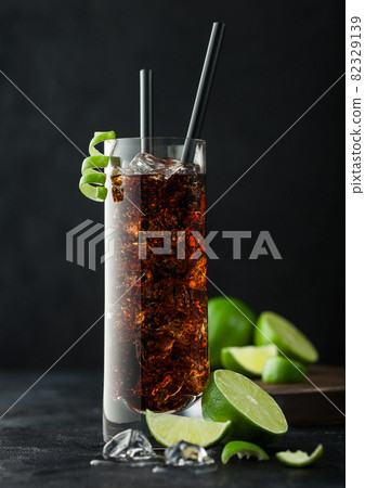 Cuba Libre cocktail in highball glass with ice and lime peel with straw and fresh limes on black background. 82329139