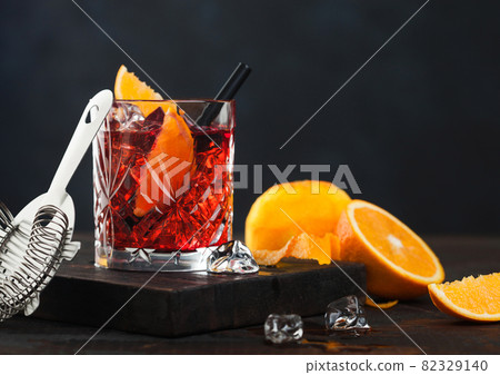 Negroni cocktail in crystal glass with orange slice and fresh raw oranges on chopping board with strainer on dark wooden background. 82329140
