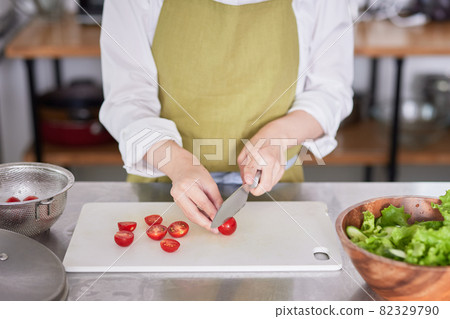 A young woman in an apron who cuts cherry tomatoes with a kitchen knife A young woman in an apron who cuts cherry tomatoes with a kitchen knife 82329790