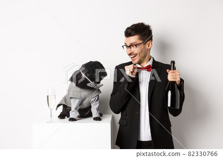 Cheerful young man in suit celebrating New Year with pet, dog and guy looking at each other, owner drinking champagne, standing over white background Cheerful young man in suit celebrating New Year with pet, dog and guy looking at each other, owner drinking champagne, standing over white background 82330766