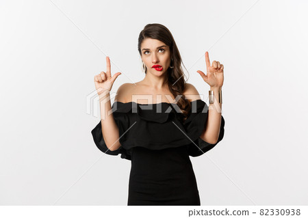 Fashion and beauty. Thoughtful attractive woman in black dress staring and pointing up, thinking with tempted look, white background Fashion and beauty. Thoughtful attractive woman in black dress staring and pointing up, thinking with tempted look, white background 82330938
