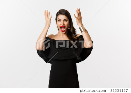 Fashion and beauty. Angry woman in black dress, shouting mad and shaking hands, grimacing outraged at camera, standing over white background Fashion and beauty. Angry woman in black dress, shouting mad and shaking hands, grimacing outraged at camera, standing over white background 82331137