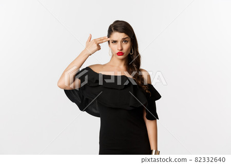 Fashion and beauty. Annoyed young woman in black dress making finger gun gesture near head, shooting herself from irritation, standing over white background 82332640