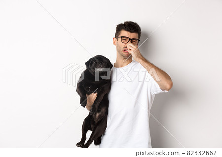Young man shut nose as holding pug, disgusted with bad smell fart of animal, standing over white background Young man shut nose as holding pug, disgusted with bad smell fart of animal, standing over white background 82332662