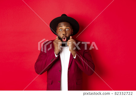 Fashion and party concept. Surprised african american male model looking shocked and amazed, saying wow and staring camera, standing against red background 82332722
