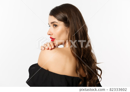 Fashion and beauty concept. Elegant woman leaning on shoulder and gazing aside with sensual piercing eyes, wearing makeup and red lipstick, standing over white background Fashion and beauty concept. Elegant woman leaning on shoulder and gazing aside with sensual piercing eyes, wearing makeup and red lipstick, standing over white background 82333602