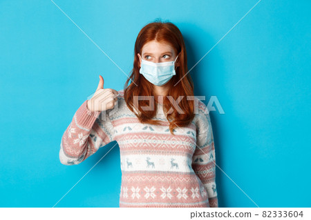 Winter, coronavirus and social distancing concept. Pretty redhead girl in face mask and sweater, looking at upper left corner promo and showing thumb up, praising good product Winter, coronavirus and social distancing concept. Pretty redhead girl in face mask and sweater, looking at upper left corner promo and showing thumb up, praising good product 82333604