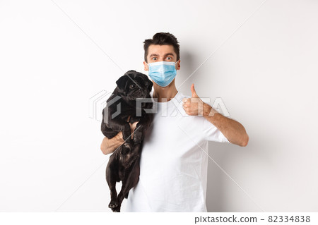 Covid-19, animals and quarantine concept. Young man in medical mask holding cute black pug dog, showing thumb up, like and approve, standing over white background Covid-19, animals and quarantine concept. Young man in medical mask holding cute black pug dog, showing thumb up, like and approve, standing over white background 82334838