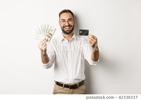 Happy young man showing his credit card and money dollars, smiling satisfied, standing over white background 82336337