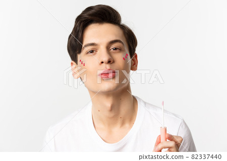 Close-up of beautiful gay man with glitter on face pucker lips after applying lip gloss, standing over white background 82337440
