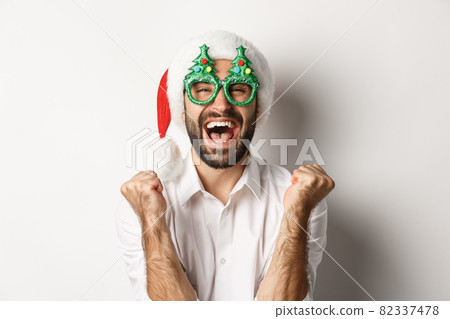Close-up of man celebrating christmas or new year, wearing xmas party glasses and santa hat, rejoicing and shouting of joy, standing over white background Close-up of man celebrating christmas or new year, wearing xmas party glasses and santa hat, rejoicing and shouting of joy, standing over white background 82337478