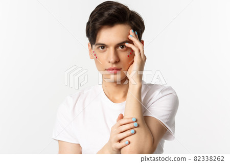 People, lgbtq and beauty concept. Close-up of beautiful queer man touching face with fingers with blue nail polish, standing over white background 82338262