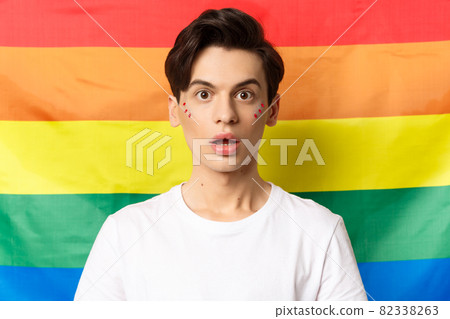 Close-up of surprised queer person with glitter on face, looking amazed at camera, standing over lgbtq rainbow flag 82338263