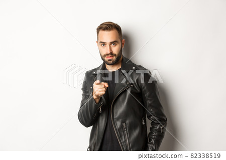 Handsome and cool bearded guy pointing finger at camera, wearing leather jacket, standing sassy against white background Handsome and cool bearded guy pointing finger at camera, wearing leather jacket, standing sassy against white background 82338519