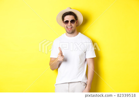 Confident and cheeky guy on vacation flirting with you, pointing finger at camera and winking, wearing summer hat with sunglasses, yellow background Confident and cheeky guy on vacation flirting with you, pointing finger at camera and winking, wearing summer hat with sunglasses, yellow background 82339518