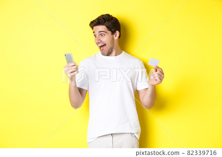 Surprised guy holding smartphone and credit card, online shopping on black friday, standing over yellow background 82339716
