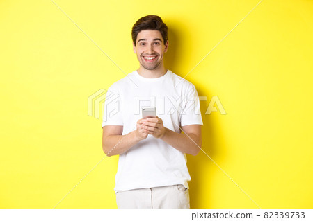 Man smiling and looking happy after reading promo offer on smartphone, standing against yellow background in white t-shirt 82339733