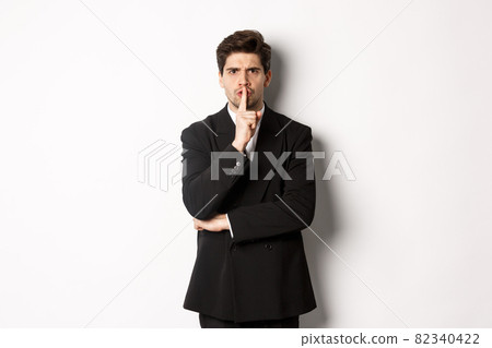 Portrait of angry boss in suit shushing at you, telling to be quiet, showing taboo hush sign and frowning, standing over white background 82340422