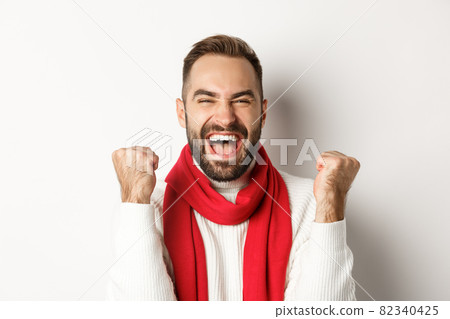 Christmas holidays. Close-up of happy guy winner, celebrating victory, raising hands up and shouting yes for joy, achieve goal, white background Christmas holidays. Close-up of happy guy winner, celebrating victory, raising hands up and shouting yes for joy, achieve goal, white background 82340425
