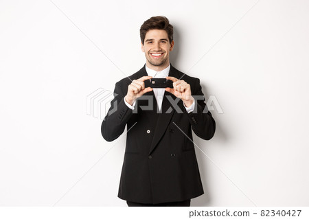 Portrait of confident handsome businessman, smiling and showing credit card, standing in suit against white background Portrait of confident handsome businessman, smiling and showing credit card, standing in suit against white background 82340427