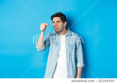 Confused guy holding something tiny in fingers and looking at it puzzled, standing over blue background Confused guy holding something tiny in fingers and looking at it puzzled, standing over blue background 82340430