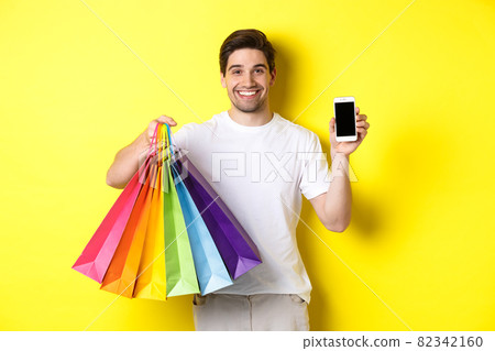 Young man holding shopping bags and showing mobile phone screen, money application, standing over yellow background 82342160