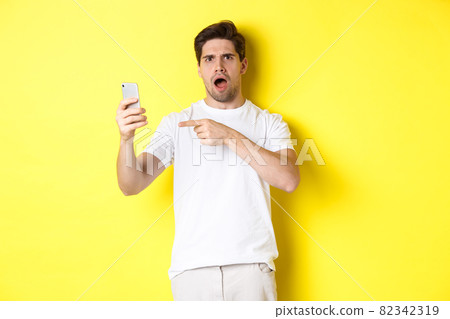Confused man pointing finger at mobile phone screen, cant understand something, standing over yellow background 82342319