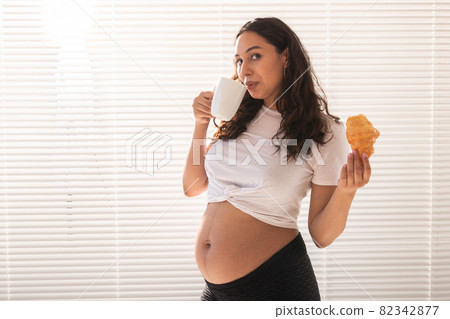 Pregnant woman eating croissant and drinks coffee. Pregnancy and maternity leave. Copy space 82342877
