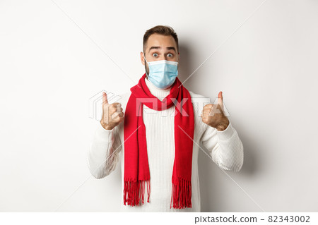 Covid-19, lockdown and quarantine concept. Surprised man in face mask showing thumbs-up, standing in sweater and red scarf, white background Covid-19, lockdown and quarantine concept. Surprised man in face mask showing thumbs-up, standing in sweater and red scarf, white background 82343002