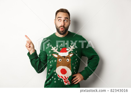 Winter holidays and christmas. Man looking curious at New Year shopping promo, standing in funny sweater against white background Winter holidays and christmas. Man looking curious at New Year shopping promo, standing in funny sweater against white background 82343016