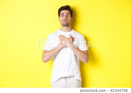 Romantic and nostalgic man holding hands on heart, close eyes and remember something, standing over yellow background Romantic and nostalgic man holding hands on heart, close eyes and remember something, standing over yellow background 82344706