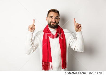 Christmas shopping and winter holidays concept. Displeased bearded guy complaining, pointing finger up and looking reluctant, standing over white background Christmas shopping and winter holidays concept. Displeased bearded guy complaining, pointing finger up and looking reluctant, standing over white background 82344709