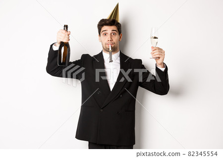 Concept of holidays and lifestyle. Handsome guy celebrating birthday, blowing party whistle and holding champagne, saying a toast, standing in suit over white background Concept of holidays and lifestyle. Handsome guy celebrating birthday, blowing party whistle and holding champagne, saying a toast, standing in suit over white background 82345554