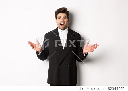 Portrait of confused and worried handsome man in suit, looking at something strange, spread hands sideways and standing puzzled against white background 82345651