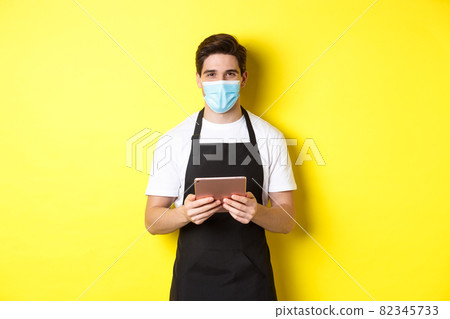 Concept of covid-19, small business and pandemic. Waiter in black apron and medical mask taking order, holding digital tablet, standing over yellow background Concept of covid-19, small business and pandemic. Waiter in black apron and medical mask taking order, holding digital tablet, standing over yellow background 82345733