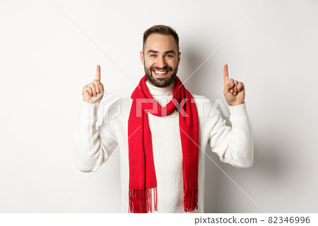 Christmas shopping and winter holidays concept. Smiling man with beard showing logo, pointing fingers up and looking happy, standing in red scarf with sweater, white background 82346996