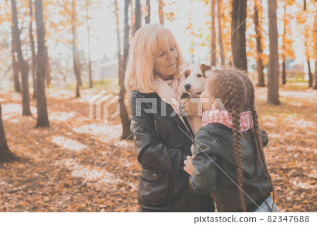 Grandmother with granddaughter in autumn park, girl hugging grandmother and her jack russell terrier dog. Generations, pet and family concept. 82347688