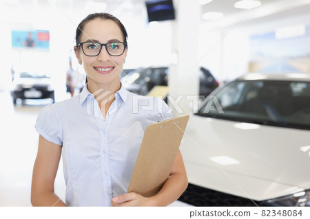 Young smiling woman manager in glasses holds clipboard on background of car dealership closeup Young smiling woman manager in glasses holds clipboard on background of car dealership closeup 82348084