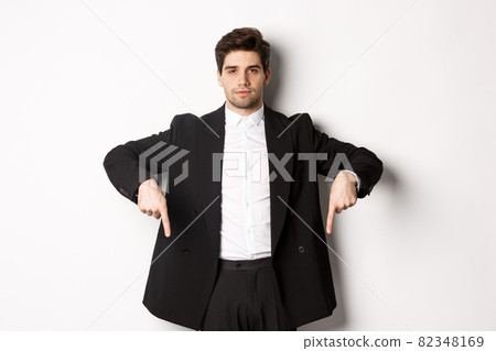 Image of handsome man dressed for formal party, wearing suit and pointing fingers down, showing advertisement or making announcement, standing over white background Image of handsome man dressed for formal party, wearing suit and pointing fingers down, showing advertisement or making announcement, standing over white background 82348169