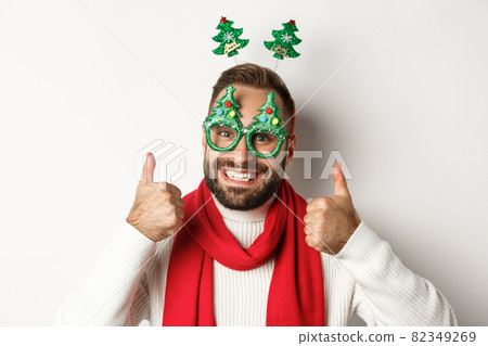 Christmas, New Year and celebration concept. Close-up of handsome bearded man in funny party glasses looking happy, showing thumb up in approval, like something, white background Christmas, New Year and celebration concept. Close-up of handsome bearded man in funny party glasses looking happy, showing thumb up in approval, like something, white background 82349269