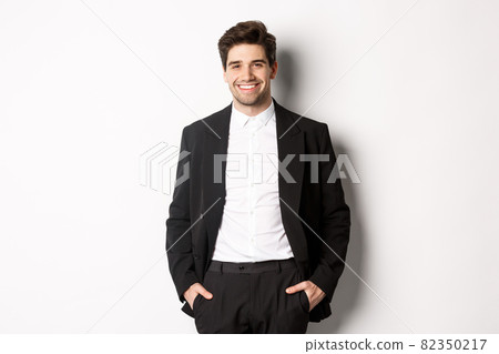 Image of handsome caucasian man in party suit, smiling pleased, attend formal event, standing over white background Image of handsome caucasian man in party suit, smiling pleased, attend formal event, standing over white background 82350217