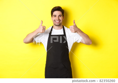 Smiling seller in black apron showing thumbs up, approve or like something, recommending cafe or store, yellow background 82350260