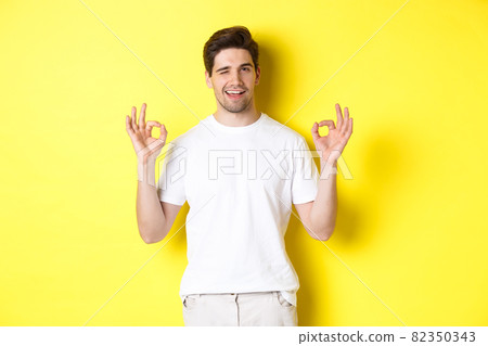 Confident handsome man winking, showing okay signs in approval, like something good, standing over yellow background Confident handsome man winking, showing okay signs in approval, like something good, standing over yellow background 82350343