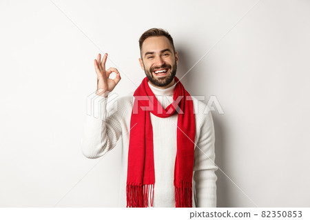 Winter holidays and shopping concept. Satisfied bearded man laughing and showing okay sign, approve and like product, standing in christmas scarf and sweater, white background Winter holidays and shopping concept. Satisfied bearded man laughing and showing okay sign, approve and like product, standing in christmas scarf and sweater, white background 82350853
