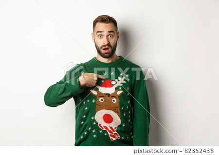 Winter holidays and christmas. Confused bearded guy pointing at himself, being startled with offer, standing over white background in sweater 82352438