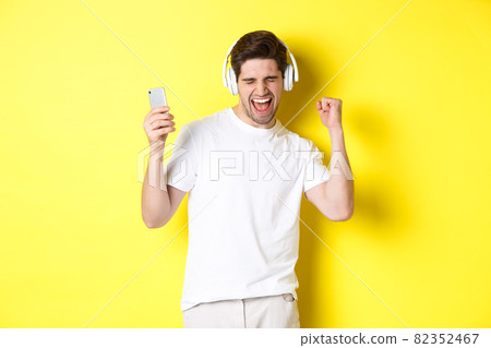 Happy man dancing and listening music in headphones, holding mobile cell phone, standing against yellow background 82352467