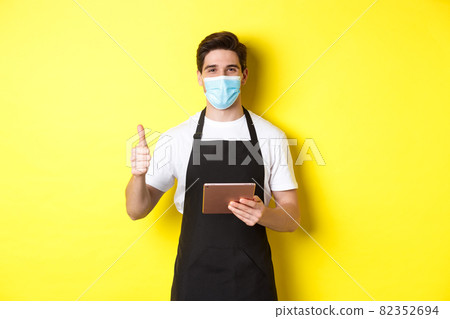 Concept of covid-19, small business and pandemic. Friendly waiter in medical mask and black apron showing thumb up, taking orders with digital tablet, yellow background Concept of covid-19, small business and pandemic. Friendly waiter in medical mask and black apron showing thumb up, taking orders with digital tablet, yellow background 82352694
