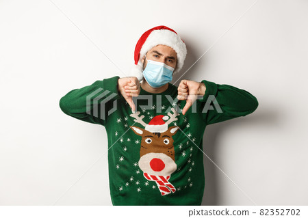 Concept of covid-19 and Christmas holidays. Displeased guy in face mask and santa hat showing thumbs down, standing over white background 82352702